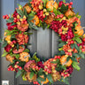 A decorative pumpkin wreath with a variety of autumn-colored flowers, including pumpkins, peonies, hydrangeas, and red berries, displayed on a door.