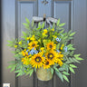 A floral wreath with yellow sunflowers, blue and yellow wildflowers, and greenery in a rustic metal bucket attached to a dark door, adorned with a black and white striped bow.