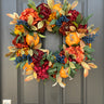 A round wreath with a autumnal design, including orange and yellow leaves, greenery, and blue berries, displayed on a dark door.